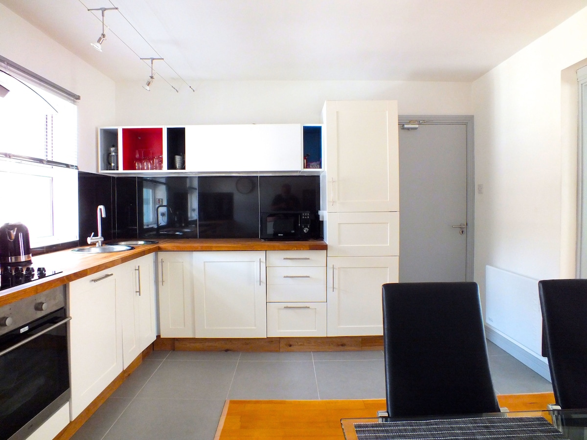 A modern kitchen is displayed, featuring sleek white cabinetry and a contrasting black backsplash. Stainless steel appliances and a wooden countertop add functionality. A dining area with four black leather chairs sits adjacent, providing a space for meals. Natural light brightens the room through a window.