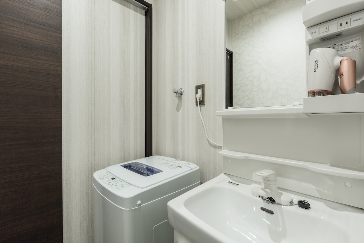 A compact bathroom area is visible, featuring a washing machine adjacent to a small sink and mirror. The walls are decorated with light floral patterns, and a hairdryer is positioned above the sink. Basic toiletry amenities are provided for convenience.