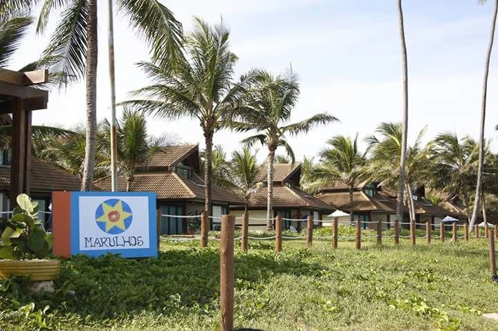 A welcoming entrance to the resort is marked by a colorful sign displaying 'Marulhos'. Lush green grass and well-maintained palm trees are seen in the foreground, providing a tropical atmosphere. Several charming buildings with tiled roofs are positioned in the background.