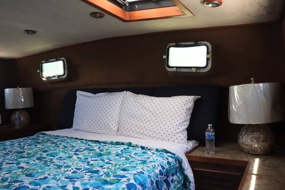 A cozy sleeping area features a spacious bed with white pillows and a colorful floral blanket. Two lamps sit on either side of the bed, and small windows are positioned above, allowing natural light to illuminate the space.