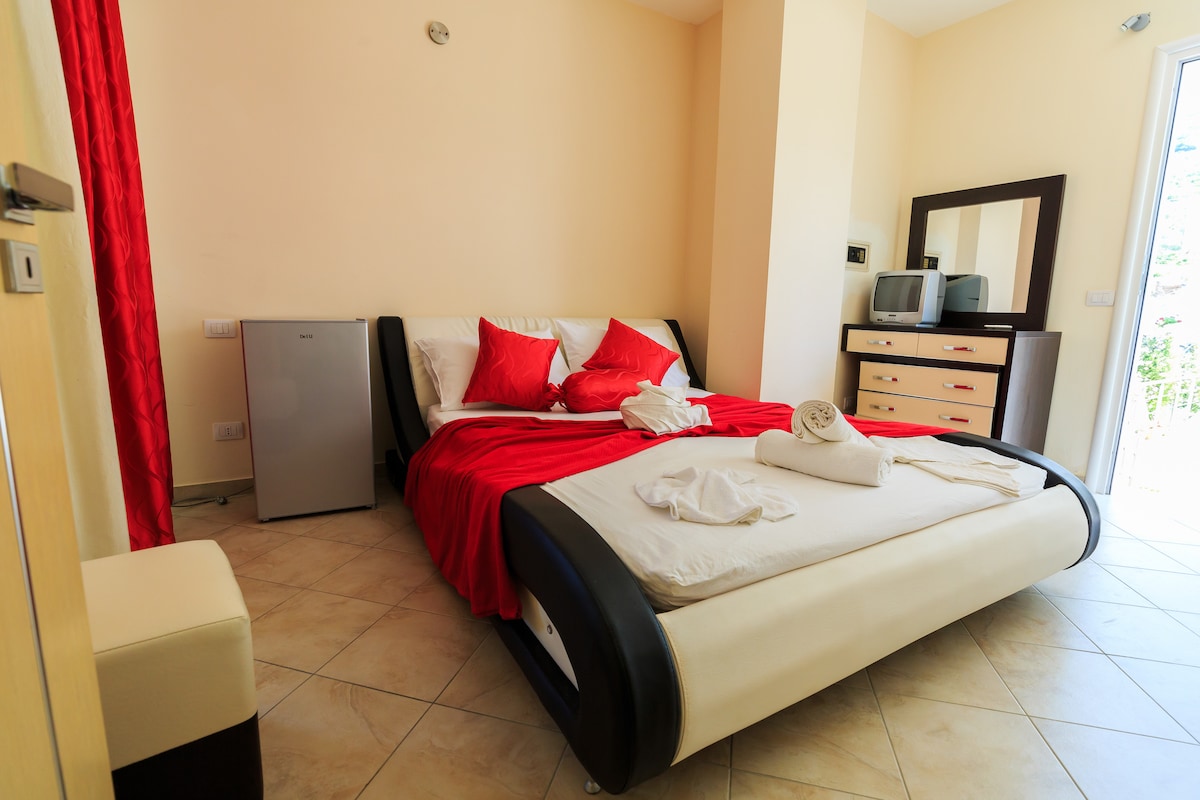 A spacious bedroom features a large bed with white linens and decorative red pillows. A mirrored dresser and a small refrigerator are positioned nearby. Natural light filters in from a glass door, leading to a private balcony with a view.