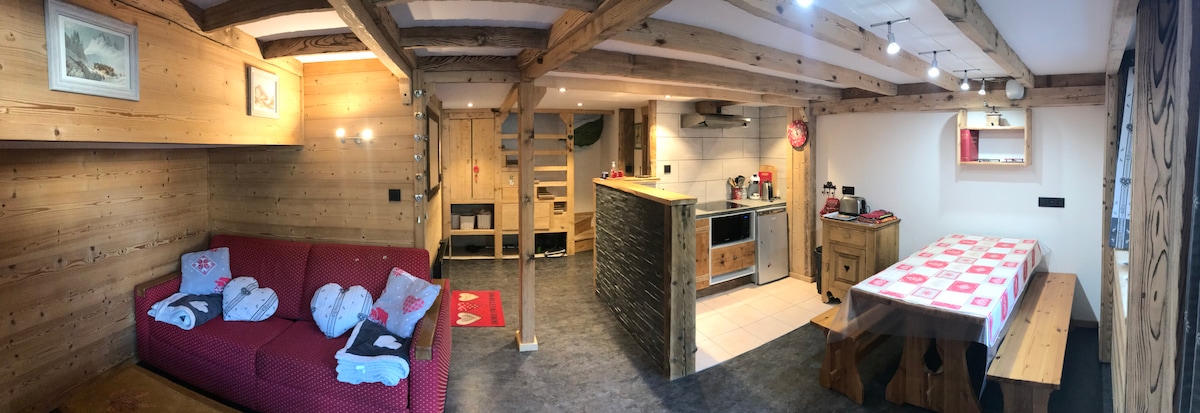 The living area features wooden beams and a cozy red sofa, complemented by a wooden dining table and benches. An open kitchen equipped with modern appliances is visible, creating a spacious layout. Soft lighting enhances the ambiance, while decorative elements add character.