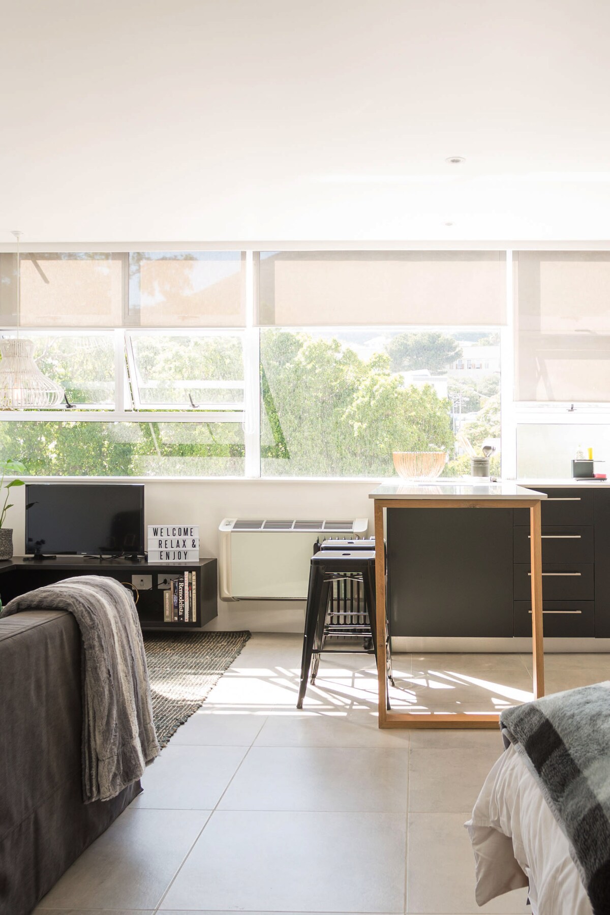 A well-lit open-plan living area features wall-to-wall windows that allow natural light to fill the space. The kitchen includes sleek cabinets and a breakfast bar, while a cozy lounge space is defined by a grey couch and decorative elements like local magazines.