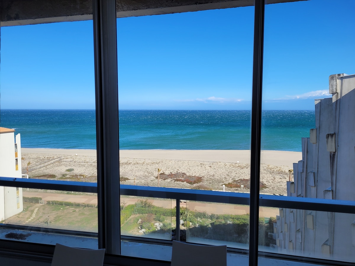 A panoramic view of the ocean can be seen through large glass windows, highlighting the clear blue water and sandy beach below. The sky is bright and open, complementing the serene coastal landscape visible from the residence.