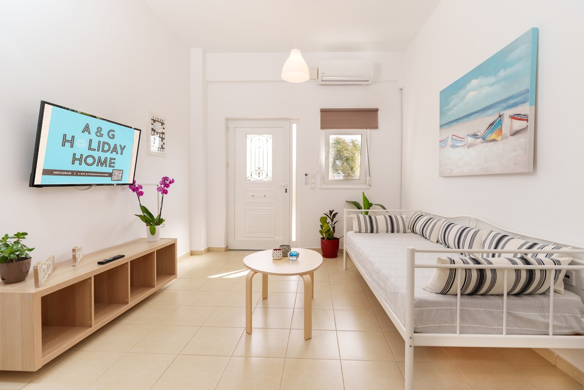 A bright living area features a white sofa adorned with striped cushions, complemented by a light wooden coffee table. A flat-screen TV is mounted on the wall, and a colorful painting of beach scenes adds a touch of charm. Natural light filters through the window.