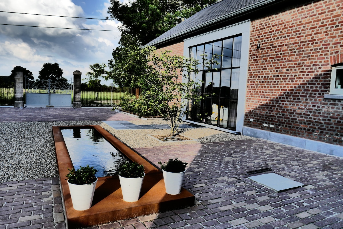 A modern, brick exterior is displayed, featuring large glass windows that reflect the surrounding greenery. A shallow water feature bordered by flower pots is visible, alongside a gravel pathway that leads to an open area with scenic views beyond.