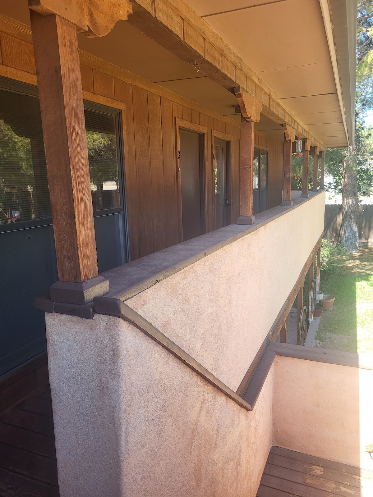 The image displays a wooden walkway leading to the entrance doors of a loft. The porch is framed by sturdy posts, and the walls feature a smooth stucco finish. Natural light illuminates the area, while a glimpse of greenery is visible in the background.