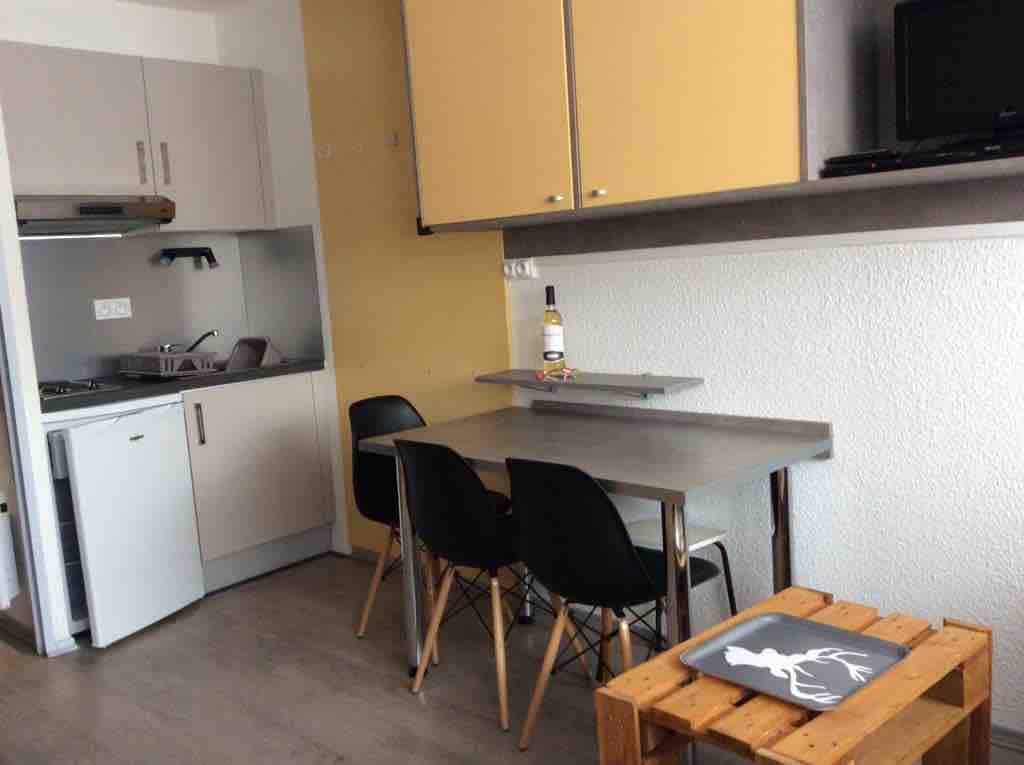 A compact kitchenette is equipped with essential appliances, including a stove and refrigerator. A small dining table with two chairs is positioned adjacent to the counter. Light-colored cabinets line the wall, while a television is mounted above the counter, adding to the functionality of the space.