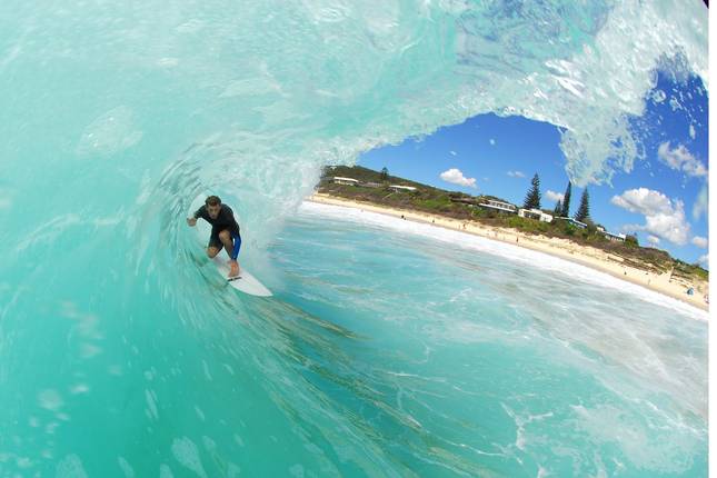 A surfer skillfully rides a wave in vibrant turquoise water. The scene captures a sandy beach in the background, with a few people visible enjoying the sun and surf. Lush greenery and a clear blue sky with scattered clouds are observed above.