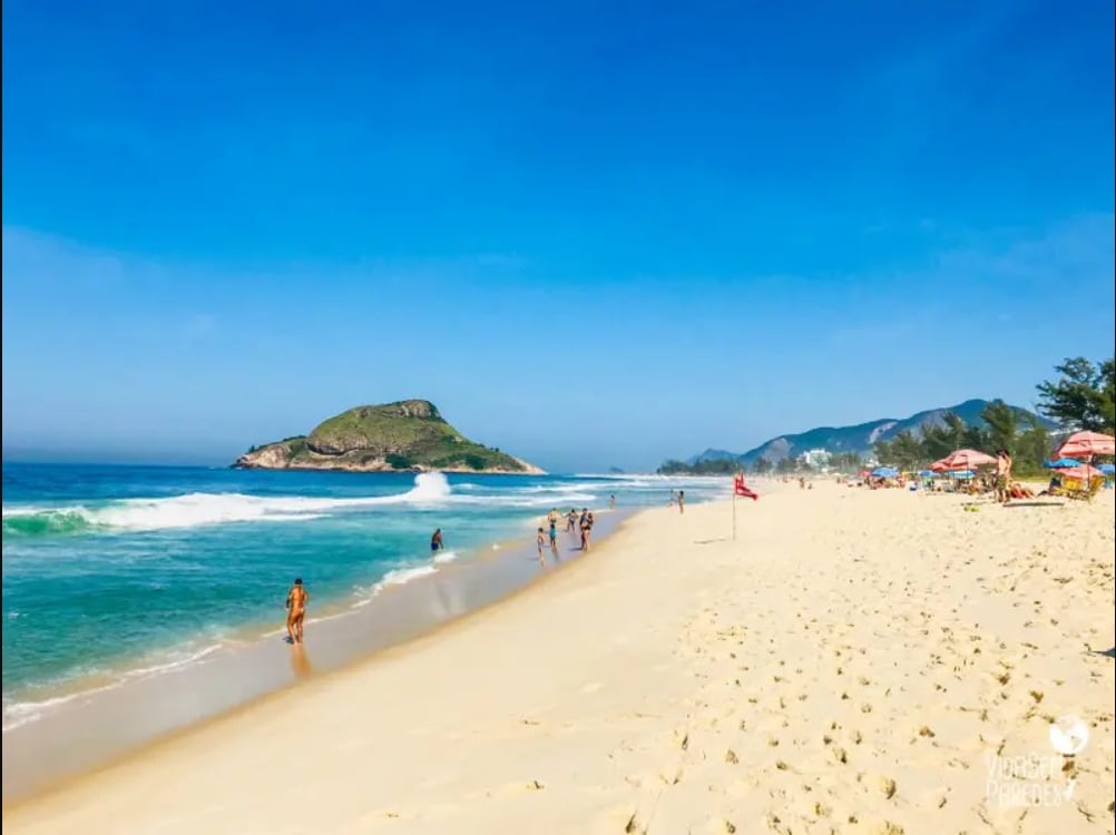 A sandy beach stretches under a clear blue sky, featuring gentle waves lapping at the shore. Small groups of people can be seen enjoying the water and sun, while colorful umbrellas provide shade in the background. Lush greenery outlines the coastal landscape.