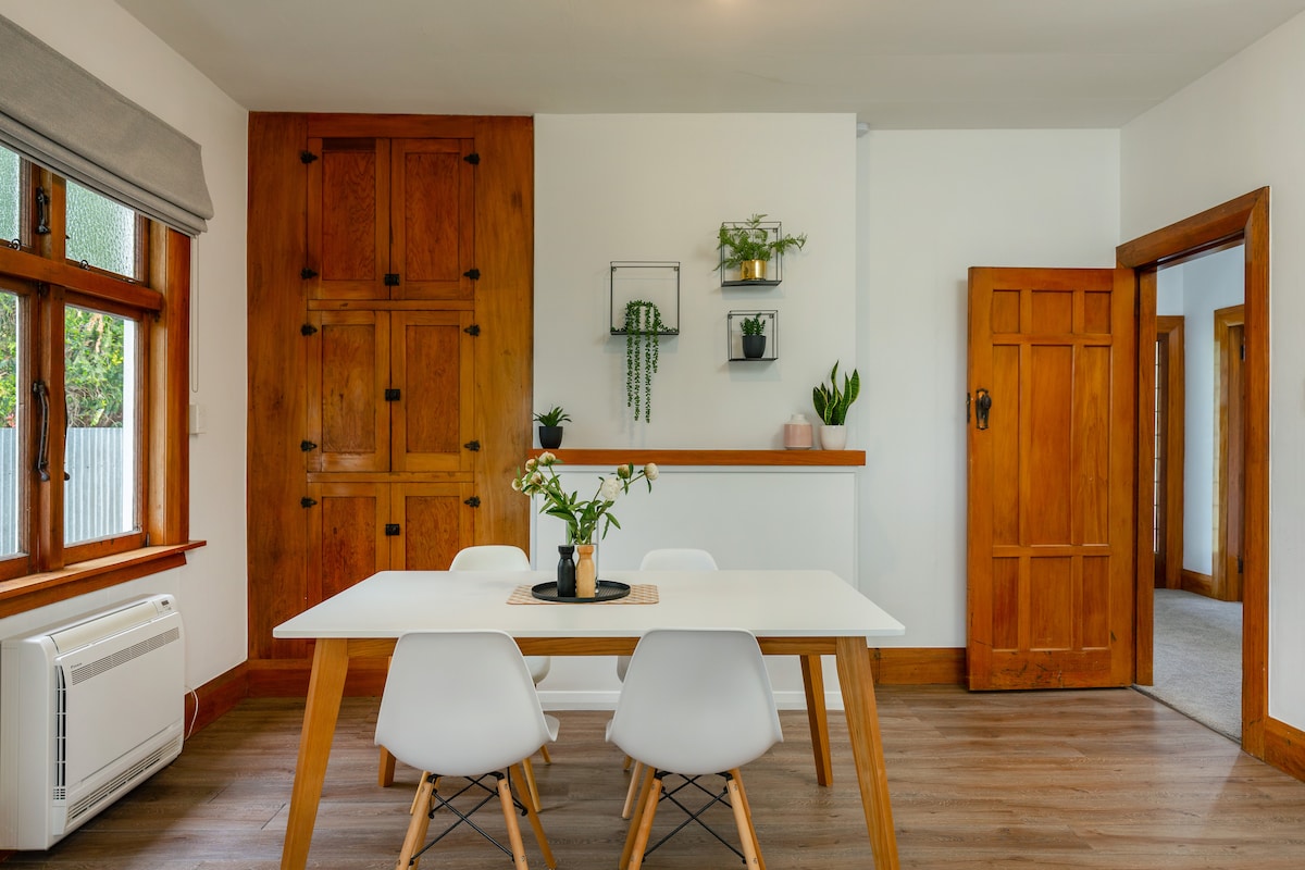 A dining area features a white table surrounded by four chairs, complemented by natural wood accents. A sideboard with decorative plants is mounted on the wall, and warm wood doors frame the entrance to another room, providing an inviting ambiance.