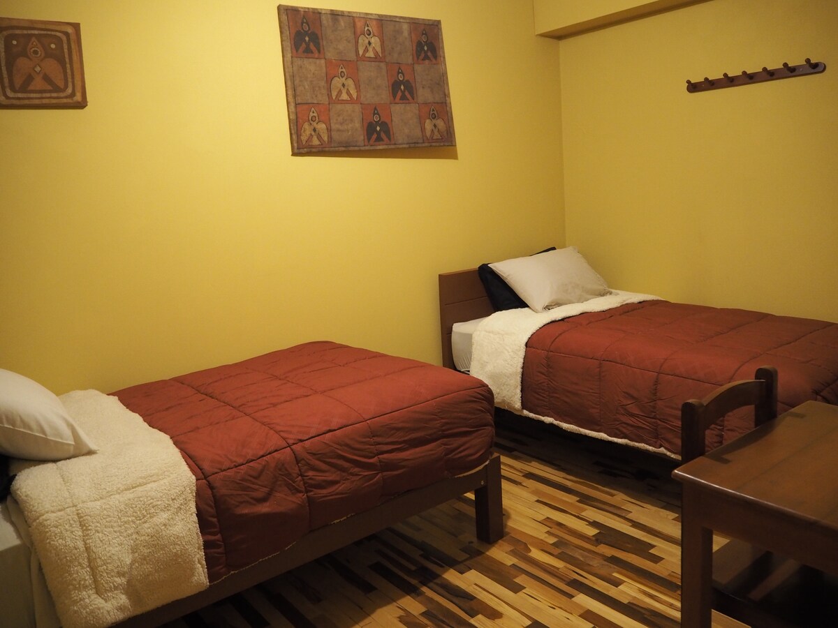 The room features two beds with warm-toned bedding, complemented by wooden flooring. A wall art piece adds character above the beds, while a small wooden table is positioned between them. Natural light fills the space, enhancing the inviting atmosphere.