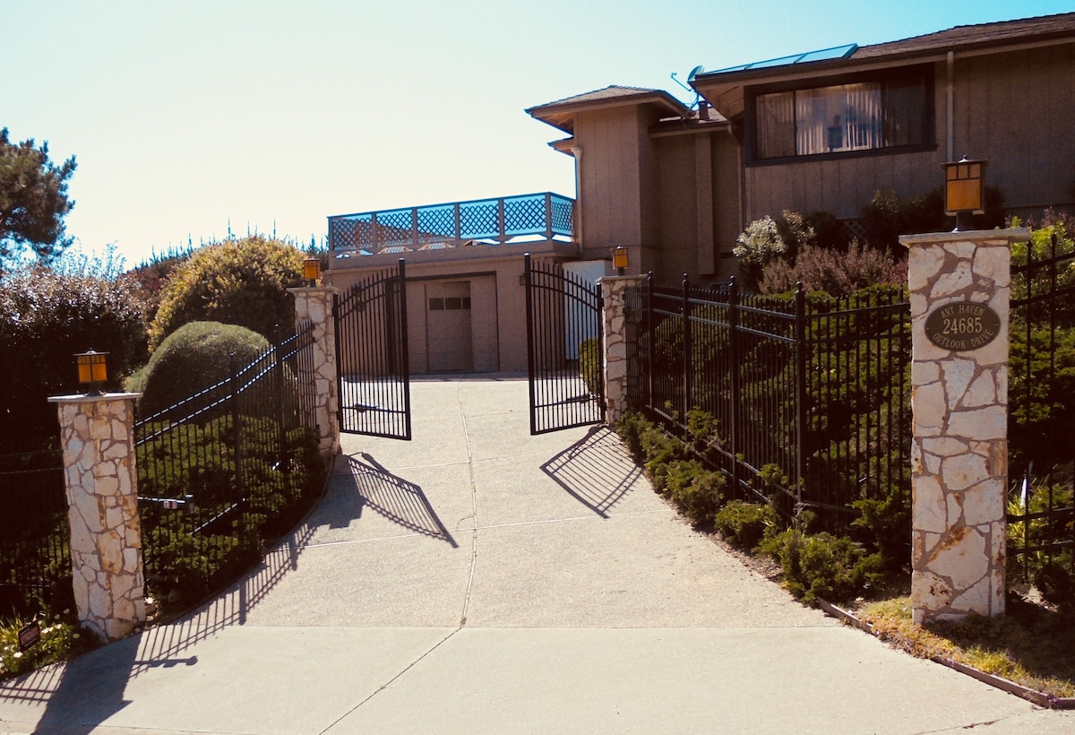 A gated entrance features decorative stone pillars and a wrought iron gate, leading up a gently sloped driveway. Lush greenery and well-maintained shrubs frame the pathway, with a two-story structure visible in the background under a clear sky.