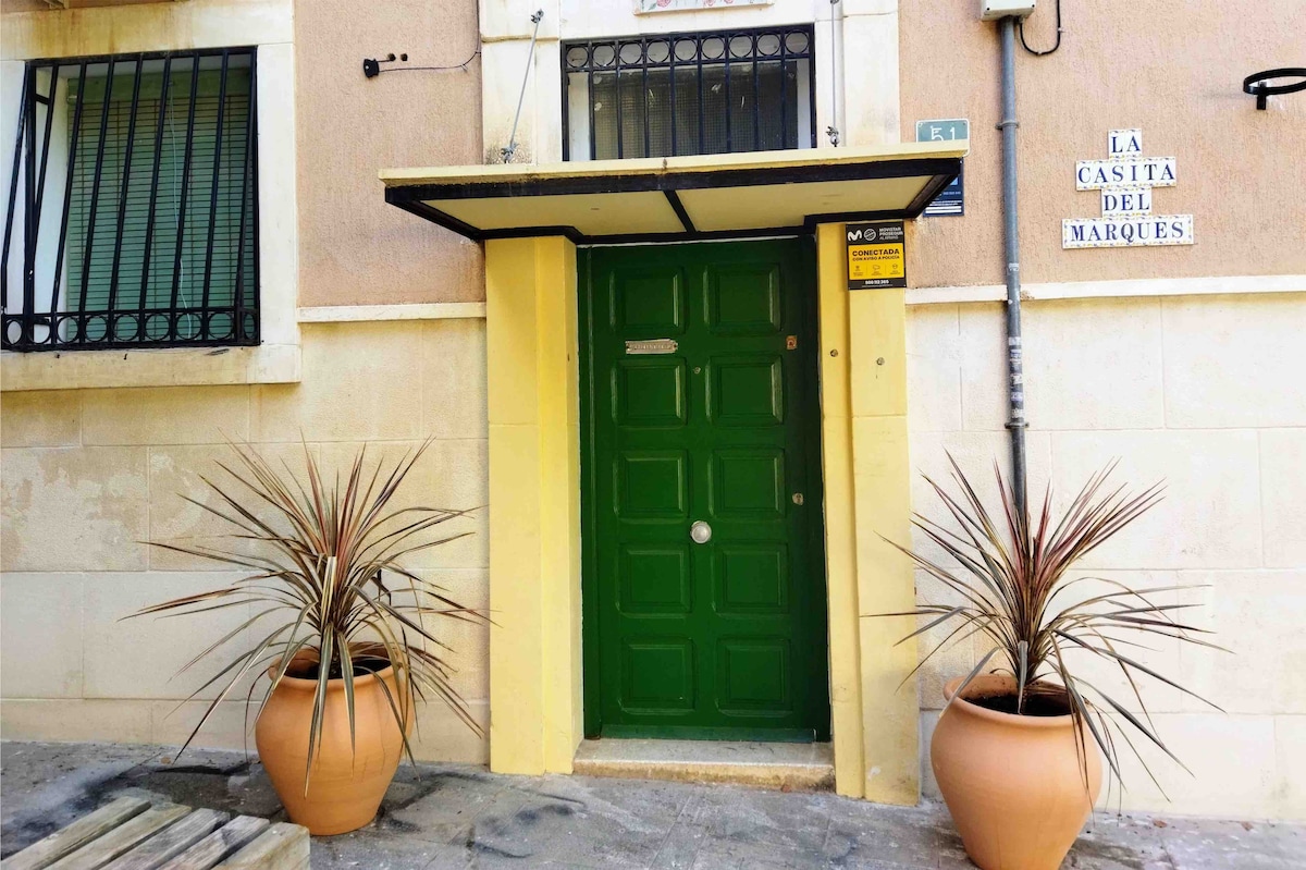 The entrance to the building is highlighted by a vibrant green door flanked by two large terracotta planters containing leafy plants. A small awning extends over the door, providing shelter. The exterior features light-colored walls and decorative signage indicating 'La Casita del Marqués.'