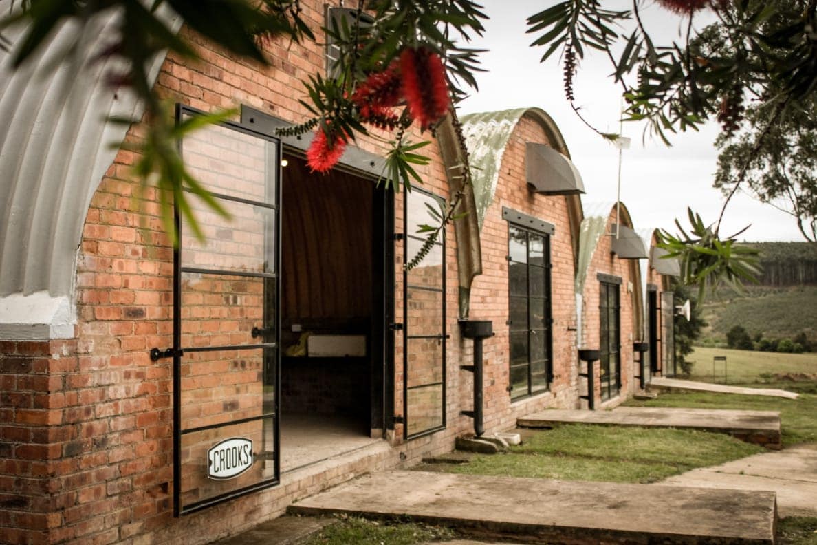 The exterior of the renovated cottage features a combination of brick and arched metal roofing. Large black-framed doors invite guests inside, with a glimpse of the lush grassy area leading to the landscaped surroundings.