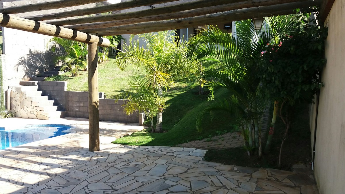 An outdoor area is showcased with a stone-paved patio shaded by a wooden pergola. Lush greenery and palms surround a swimming pool, with grassy slopes and landscaped steps leading to the residence. The space offers a serene environment ideal for relaxation.