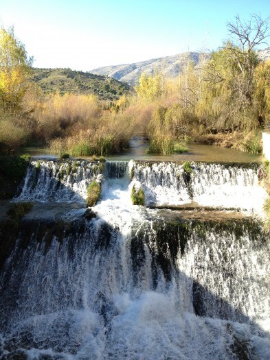 A gentle waterfall cascades down rocky steps, surrounded by lush greenery and autumn foliage. The landscape features distant mountains, framing the scene under a clear blue sky. Tranquil waters flow in the foreground, reflecting the natural beauty of the serene environment.