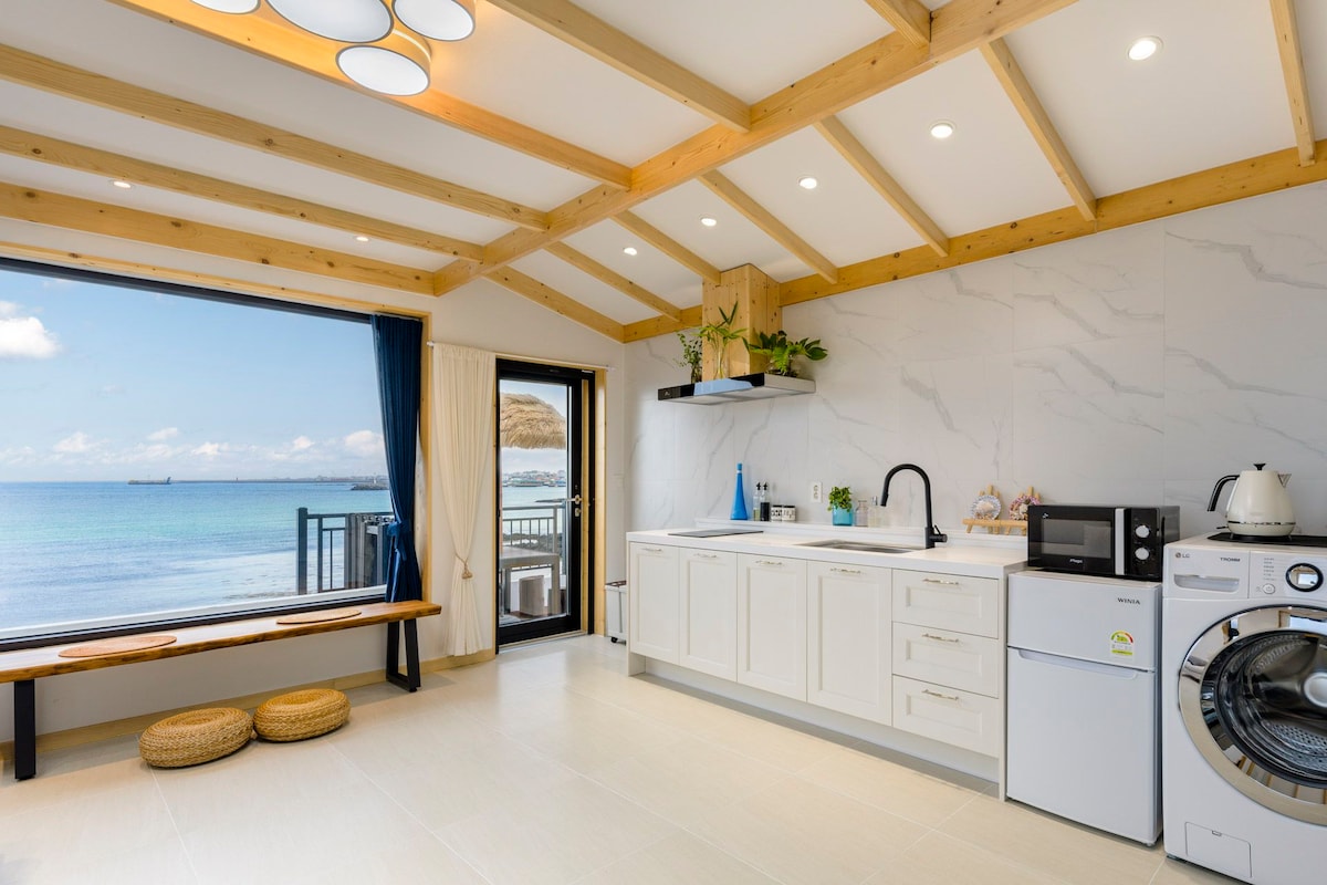 A bright kitchen area features white cabinetry and modern appliances. Large windows provide views of the sea, allowing natural light to fill the space. Wooden beams on the ceiling add warmth. A seating area with cushions is visible near the window.