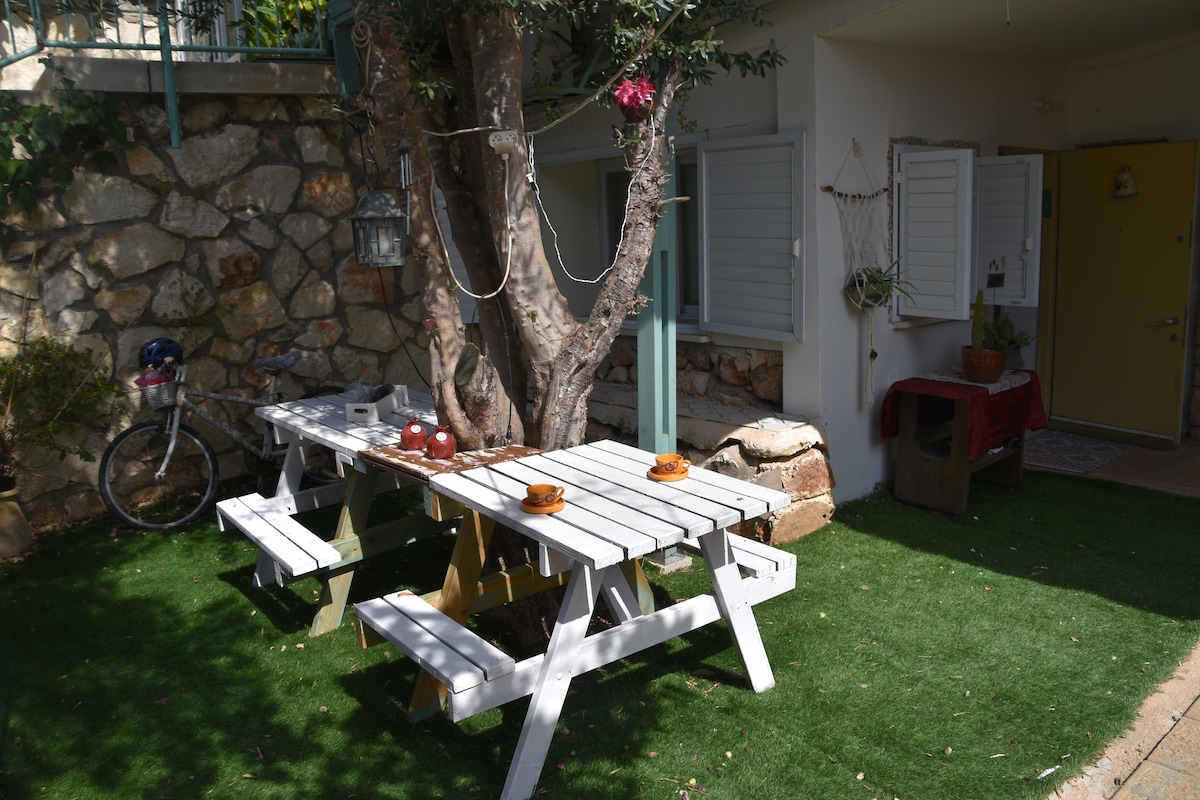 A charming outdoor picnic area features a large wooden table with attached benches, set on a grassy lawn. An olive tree provides shade, while a stone wall creates a rustic backdrop. A bicycle rests nearby, and a cozy seating nook is visible through the open windows.