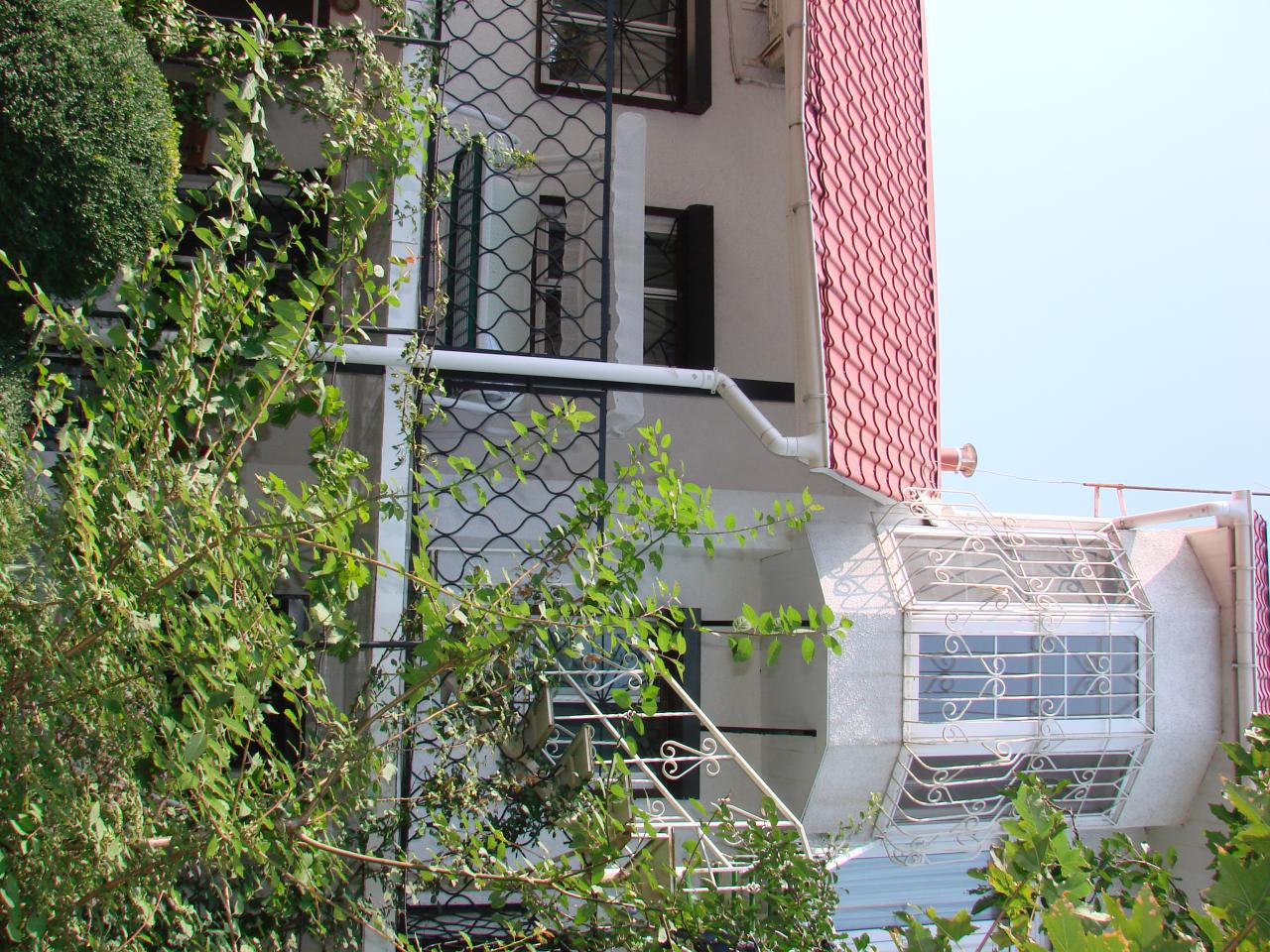 The exterior of a three-story house is visible, featuring a distinctive turret-like design with large windows. A red-tiled roof complements the white walls, while greenery surrounds the property, enhancing its private feel. A balcony with decorative railings is seen on the second floor.