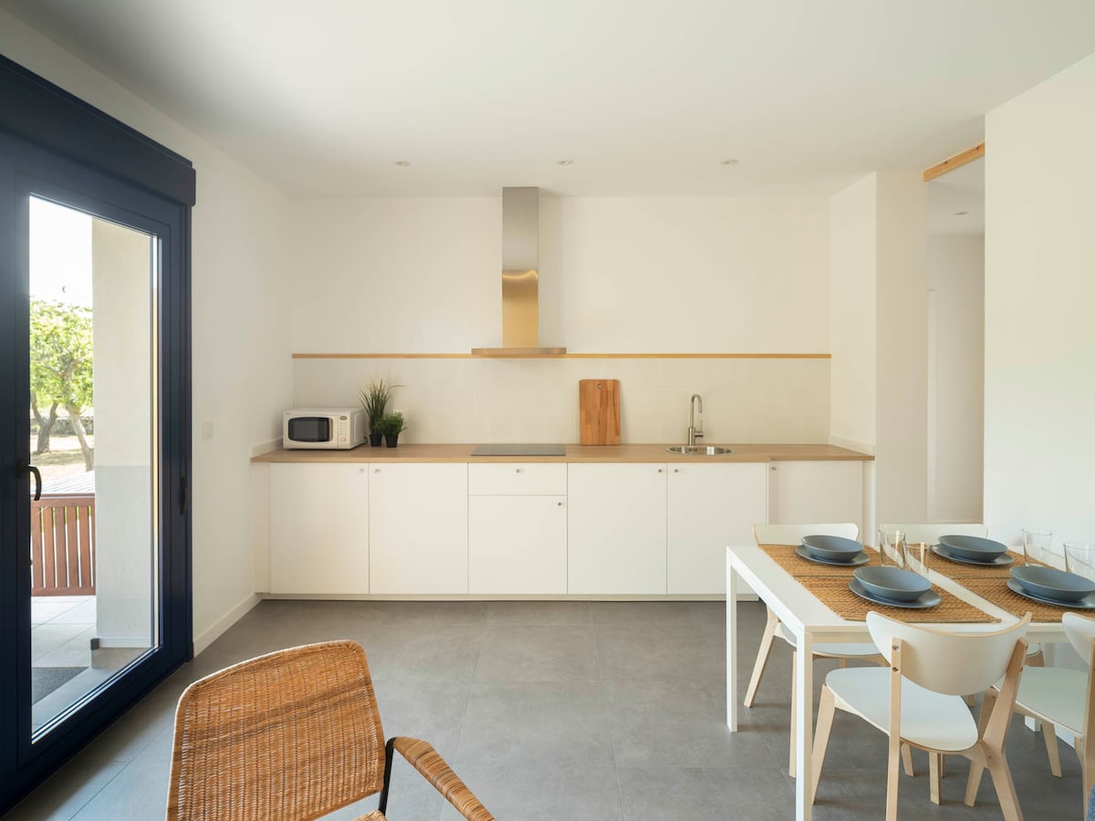 A modern kitchen features white cabinetry and a wooden countertop. A sink and stove are integrated into the design, with a microwave and a cutting board visible. A dining table is set for six with plates and cutlery, complemented by woven placemats. Natural light enters through sliding glass doors.