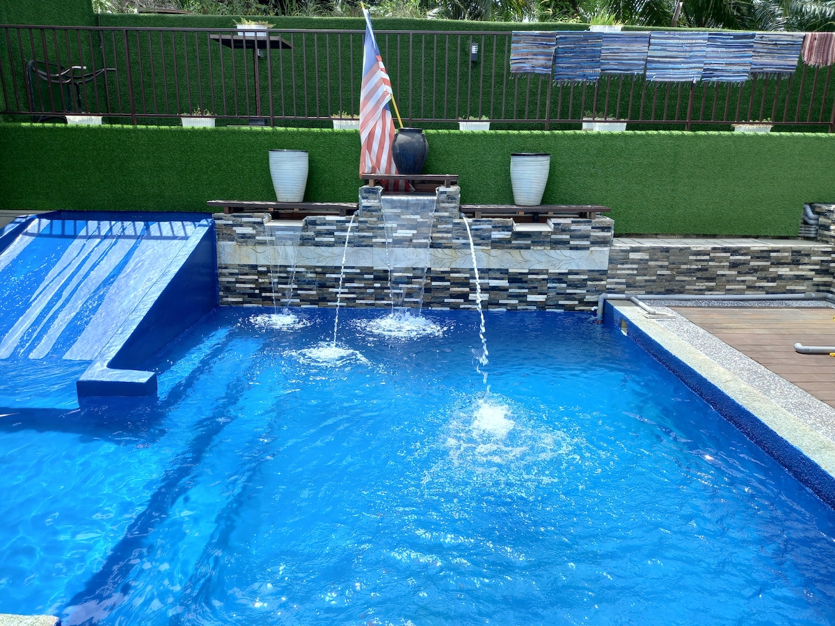 A refreshing swimming pool features a gentle waterfall and a water slide. The vibrant blue water is surrounded by stone accents and green decorative grass. Potted plants and a flag are visible along the fence, creating a relaxed outdoor space.