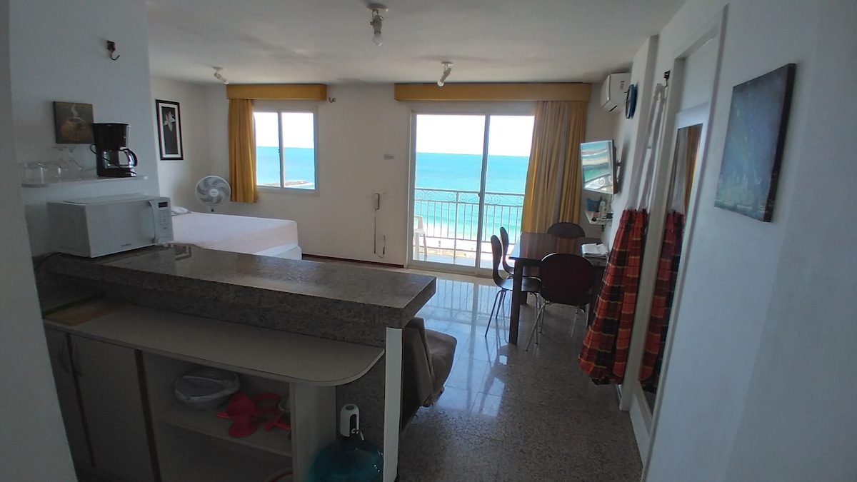 An open living space features a compact kitchen area with a breakfast bar and seating. Large windows offer a view of the ocean, complemented by light curtains. A dining table is positioned beside a mirror, enhancing the natural light within the space.