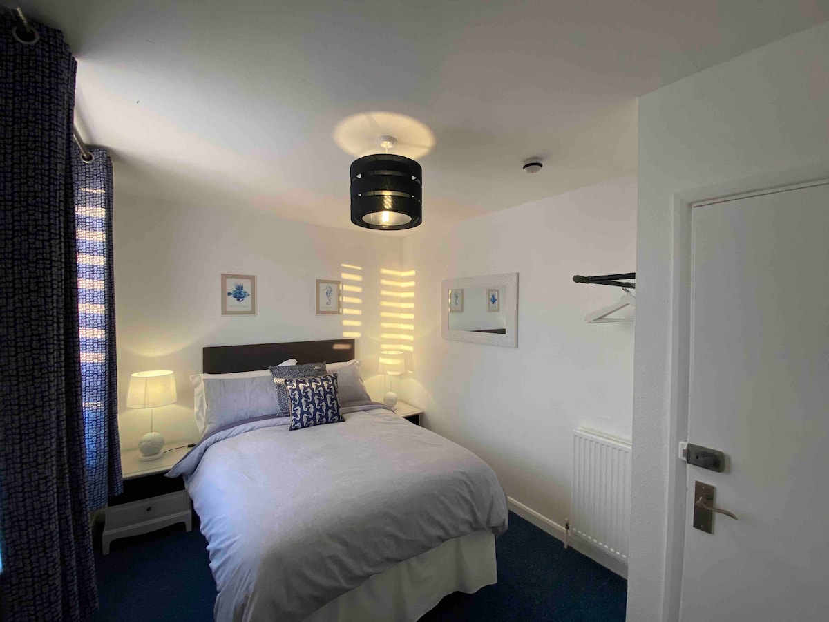 A well-lit bedroom features a contemporary design, including a spacious bed with neutral bedding and decorative pillows. Light fixtures and bedside tables enhance the inviting atmosphere. A wardrobe and hanging space are present, with natural light filtering through patterned curtains.
