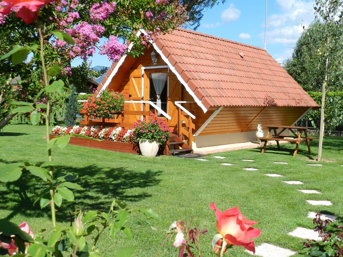 A charming wooden chalet features a distinctive red-tiled roof, surrounded by vibrant flower beds. The well-maintained lawn showcases lush greenery, and a stone path leads to a picnic table. Flowering plants add color and warmth to the picturesque setting.