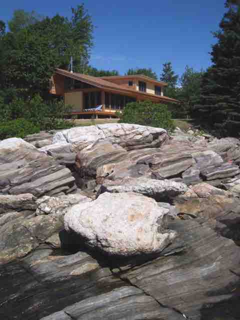 A charming cottage rests on elevated wooden decking, surrounded by a natural landscape. Below, the rugged rocks are partially covered by water, reflecting the serene blue sky. Lush greenery frames the scene, creating a tranquil outdoor setting by the coastline.