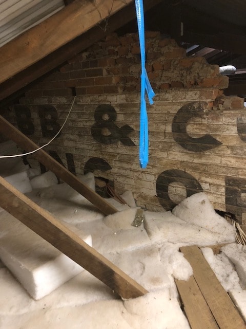 Exposed brick walls reveal old signage from the Cobb and Co staging station. Weathered wooden beams provide structure above a floor covered in insulation material. Natural light enters from a small opening, illuminating elements of the historic architecture.