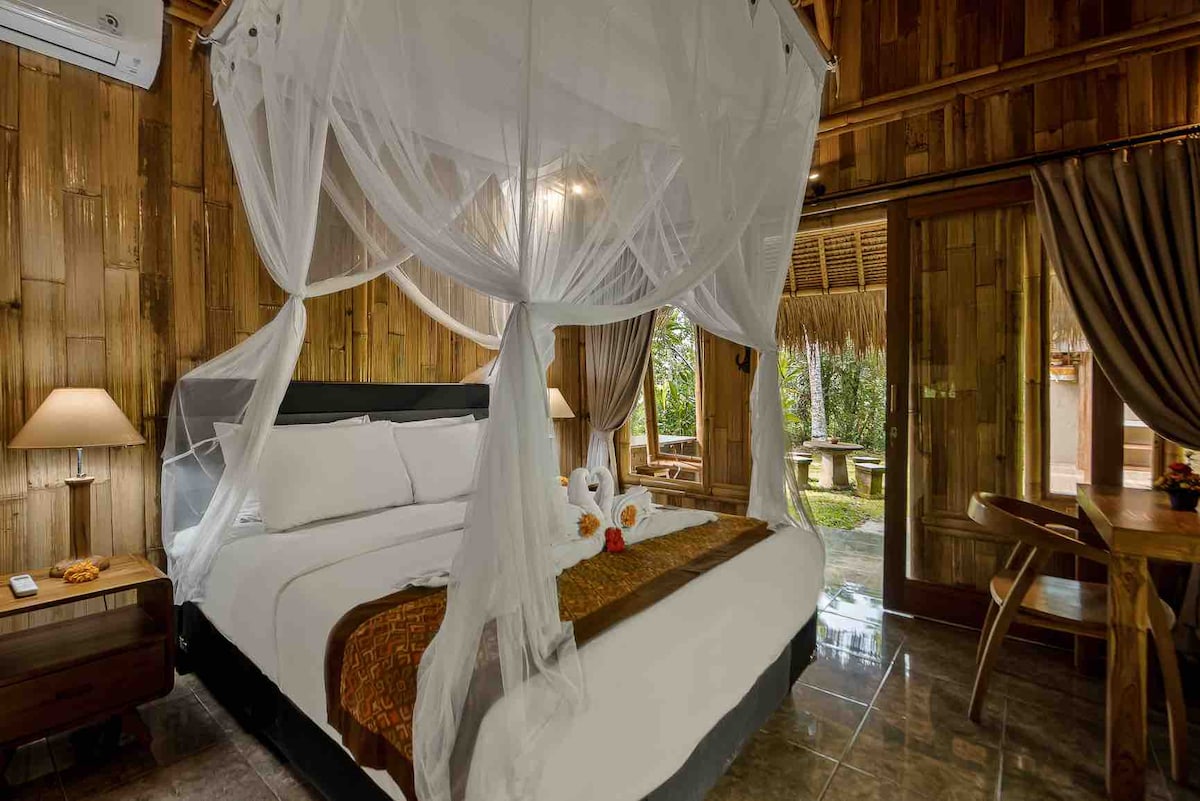 A spacious bedroom is enveloped in natural wood paneling, featuring a large bed dressed in white linens and topped with decorative accents. Soft lighting from bedside lamps creates a cozy ambiance. Sliding glass doors open to a view of lush greenery.