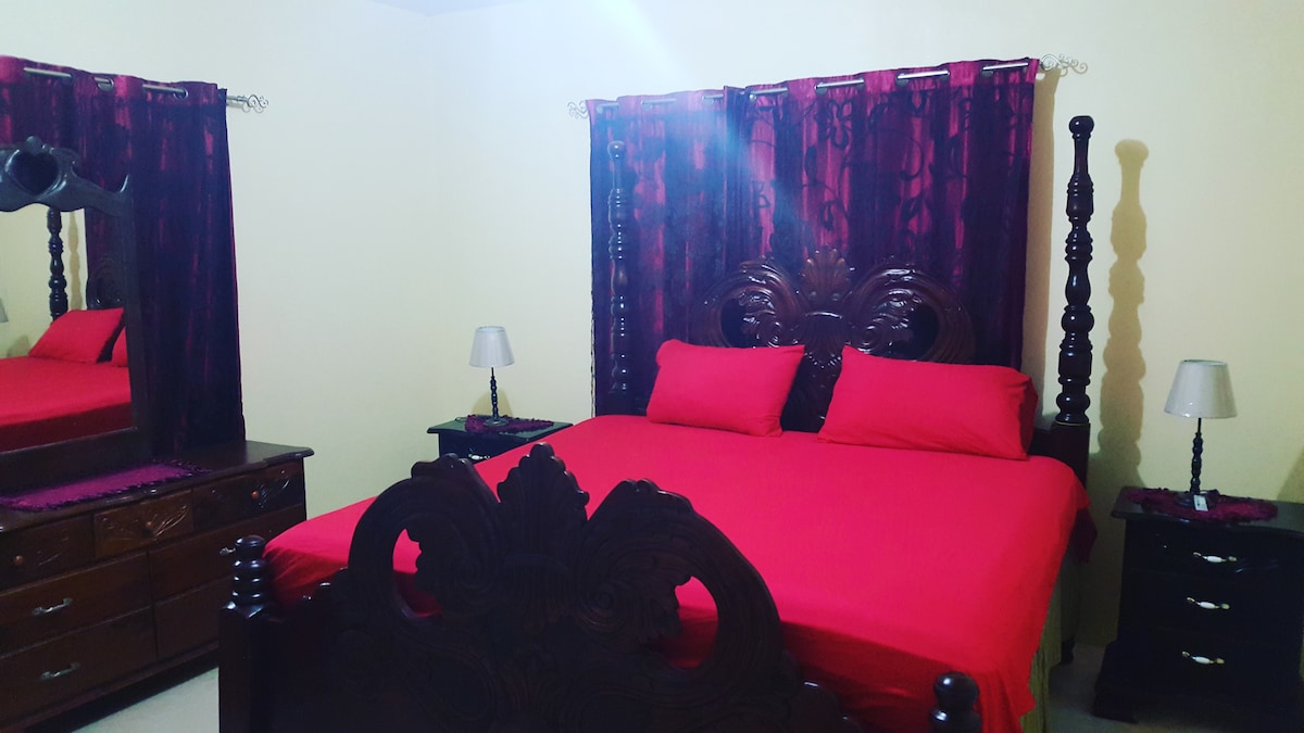A spacious bedroom features a large wooden bed with a decorative headboard, draped in bright red bedding. Two bedside tables with lamps are positioned on either side of the bed. A matching dresser reflects the room's furnishings, while dark curtains frame a window.