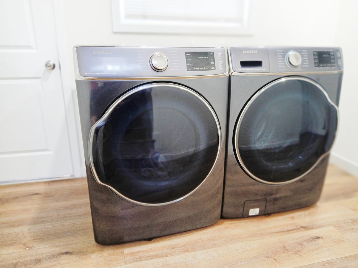 A set of modern front-loading washer and dryer units are positioned side by side against a light-colored wall. The appliances display a sleek design in a dark finish, with control panels featuring digital displays and buttons, complementing the light wood flooring.