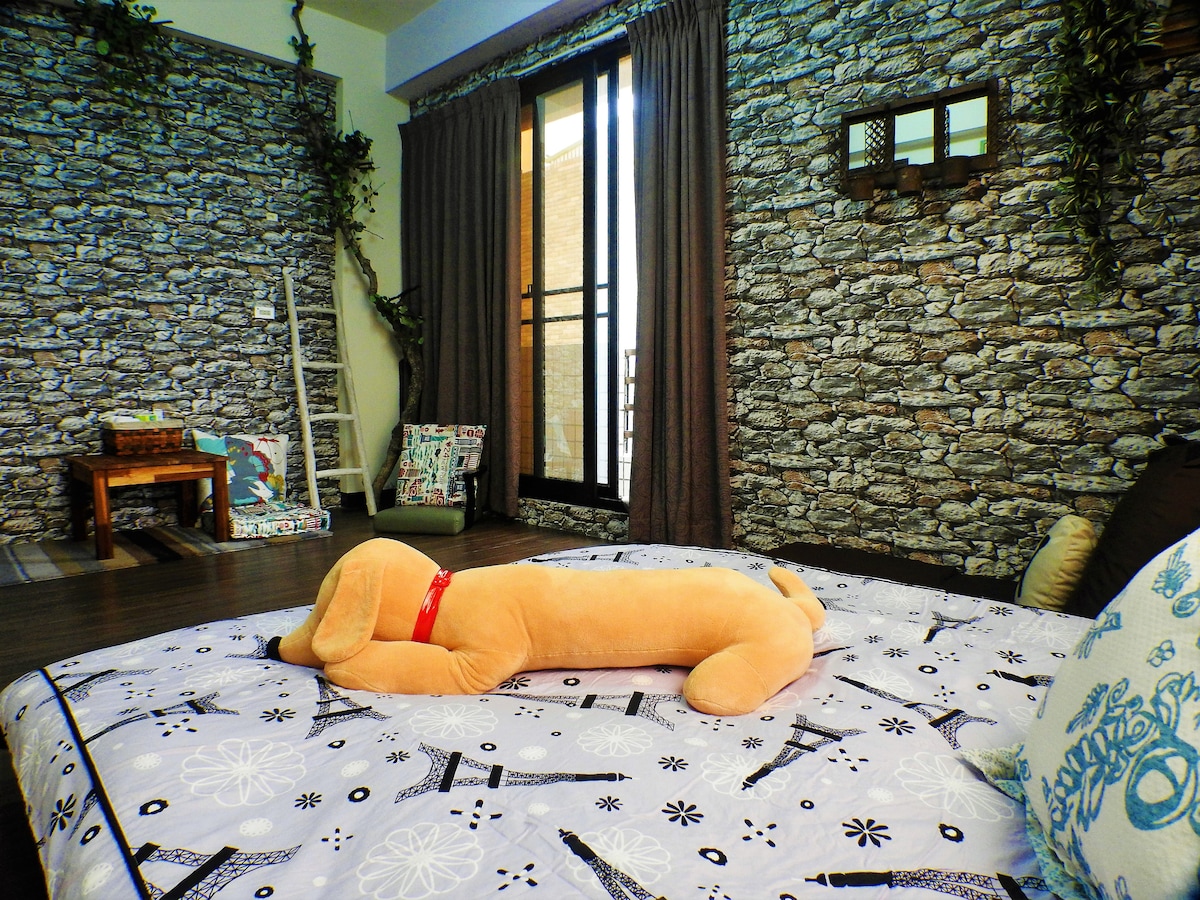 A cozy sleeping area features a spacious bed with a patterned blanket. Against a backdrop of textured stone wallpaper, a plush toy is positioned comfortably on the bed. Natural light filters through large windows, complemented by soft curtains and decorative cushions scattered around.