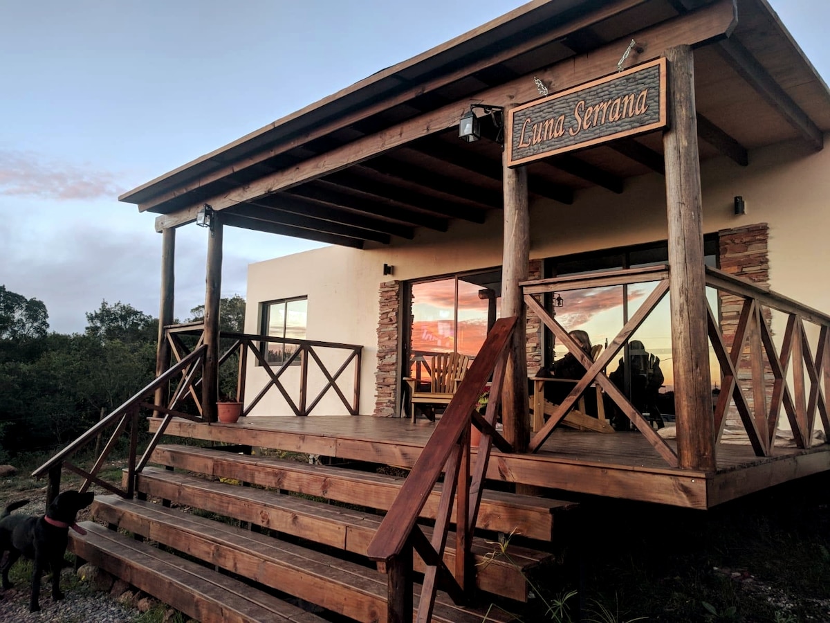 A wooden structure with a welcoming sign rests on a hillside, featuring a spacious porch with railing. Large glass doors reflect the surrounding landscape, while a set of steps leads to the entrance. Natural elements create an inviting connection to the outdoors.