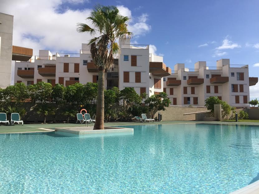 A serene pool area is featured with clear turquoise waters surrounded by greenery. Comfortable lounge chairs are positioned along the water's edge. The backdrop includes a modern residential building with prominent balconies and light-colored façades, set against a clear blue sky.