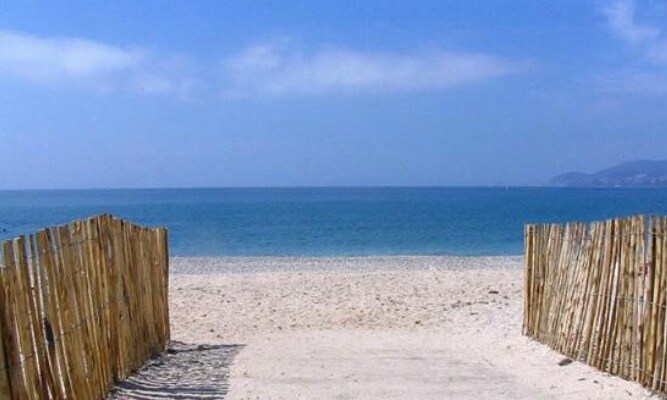 A sandy pathway leads to a calm beach, flanked by a simple wooden fence. The clear blue sea stretches out to the horizon under a bright sky, creating a serene coastal view.