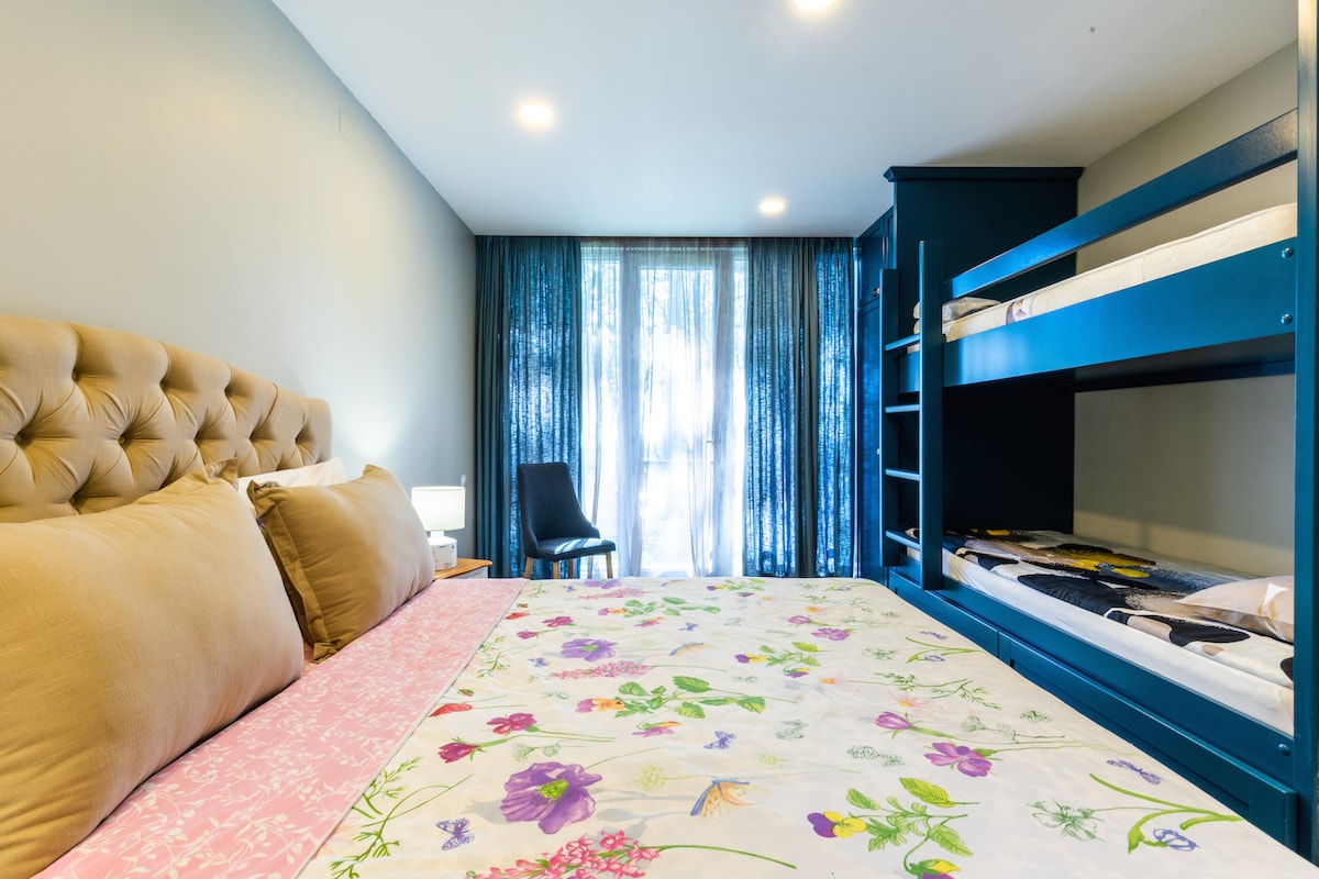 A multi-functional bedroom features a spacious bed adorned with a floral bedspread. A built-in bunk bed is positioned against a blue wall, with light filtering through sheer curtains that frame a window. A chair is placed nearby, adding to the room's practicality.