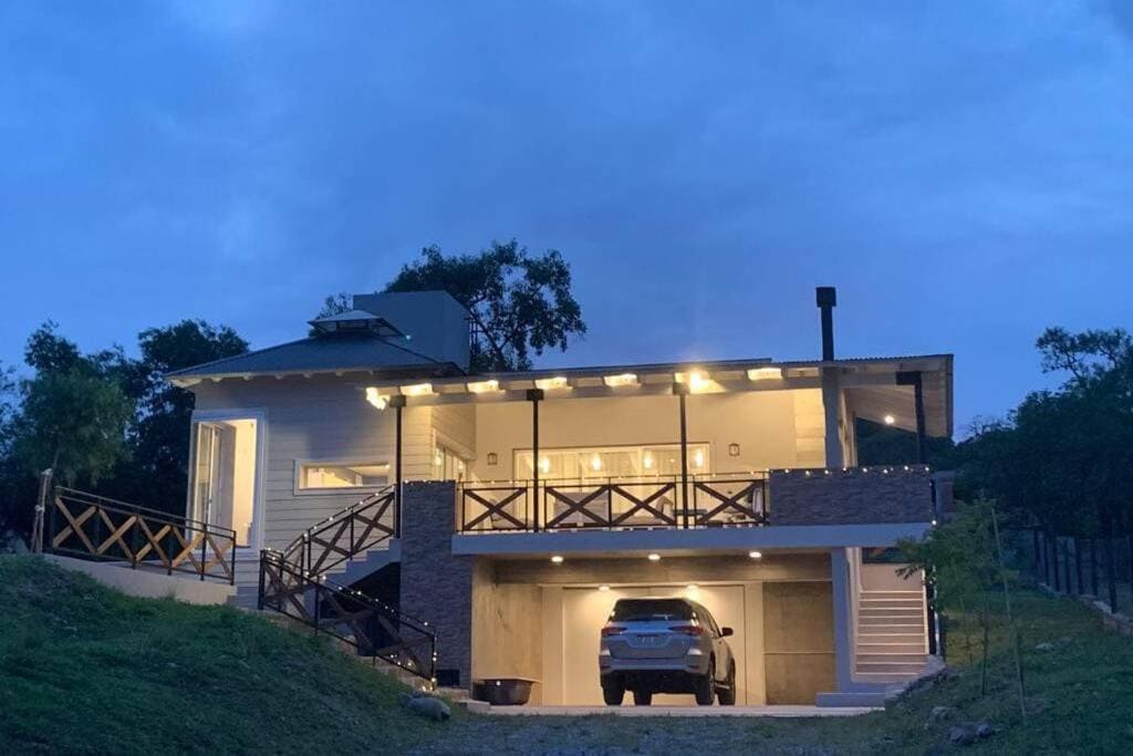 A modern house is showcased in the twilight, featuring a spacious balcony with wooden railings. The exterior is complemented by soft lighting, which highlights the entrance and supports a relaxed ambiance. A car is parked in the designated space beneath, surrounded by lush greenery.