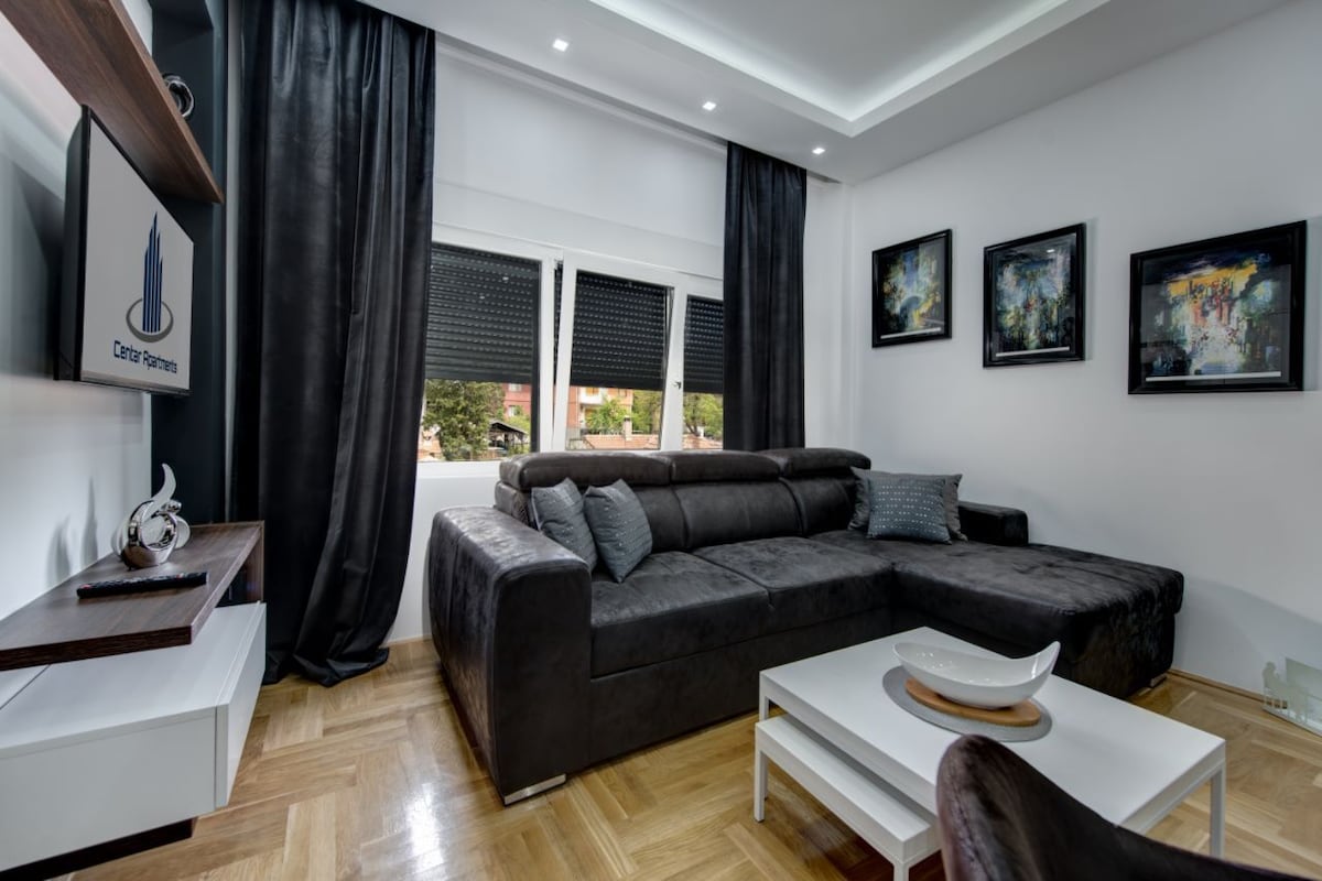 A modern living area features a dark sectional sofa positioned near large windows covered with practical blinds. Four art pieces are displayed on the walls, and a sleek white coffee table rests on a polished wooden floor. Soft lighting contributes to a cozy ambiance.