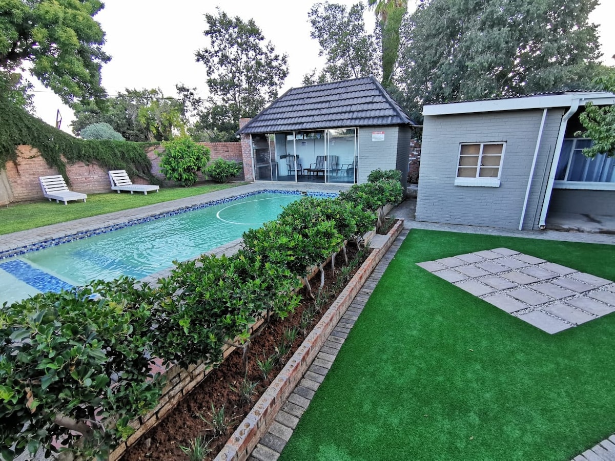 An outdoor area is showcased with a sparkling swimming pool bordered by neatly trimmed hedges. Sun loungers are arranged by the pool, and a grassy lawn complements the space. A modern structure with large glass doors can be seen, providing a view of the inviting yard.