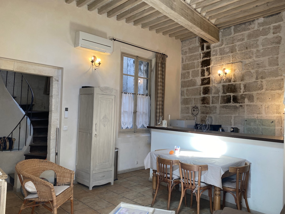 A spacious living area features a light-coloured dining table surrounded by four wooden chairs. Natural light enters through a window with sheer curtains, illuminating the room's stone walls and wooden beams. A white cabinet is positioned beside the bar area, creating an open feel.
