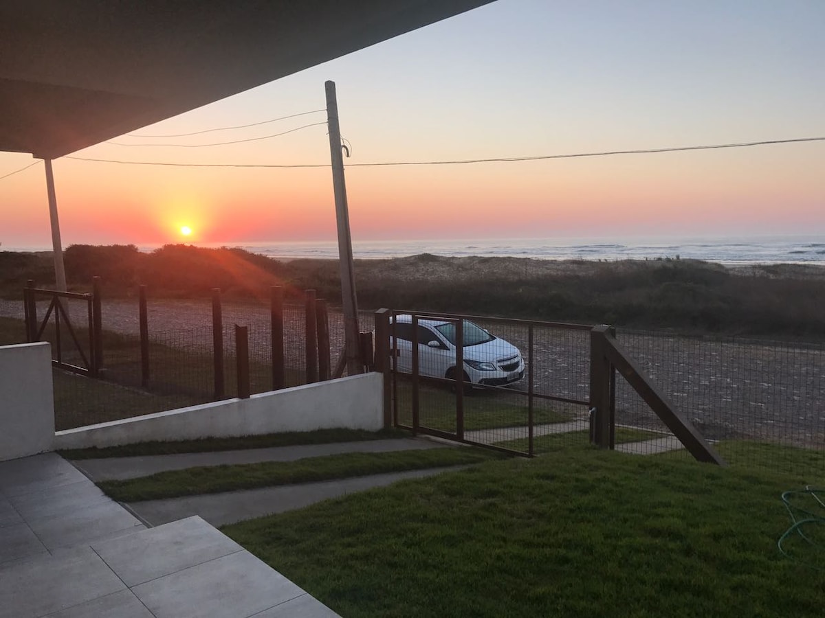 A serene view showcases a colorful sunrise over the ocean, framed by a grassy area and a pathway leading to the beach. A car is parked near a fenced area, with the gentle waves visible in the distance.