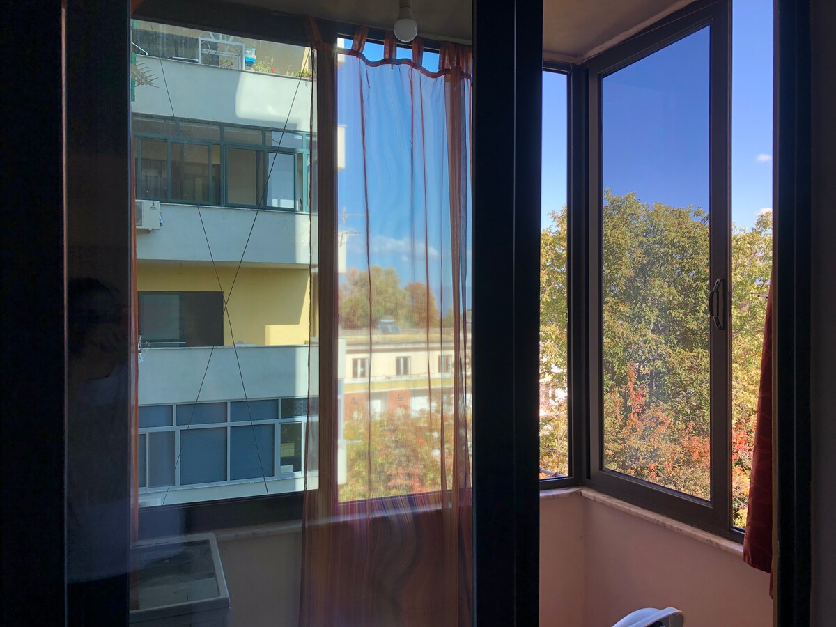 A balcony space is visible, featuring large windows that allow natural light to fill the area. Sheer curtains hang gently, filtering sunlight and offering glimpses of vibrant autumn foliage outside. The backdrop includes a neighboring building and a clear blue sky.