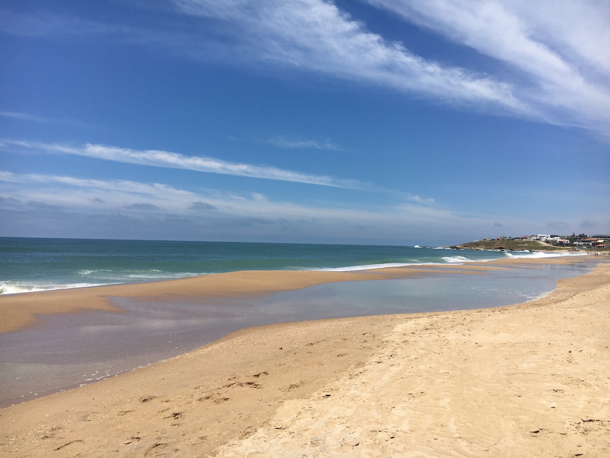 An unobstructed view of a sandy beach stretches along the coastline, where gentle waves meet the shore. The sky is clear with a mix of soft clouds. The tranquil scene conveys a sense of openness and natural beauty, providing an ideal setting for relaxation.