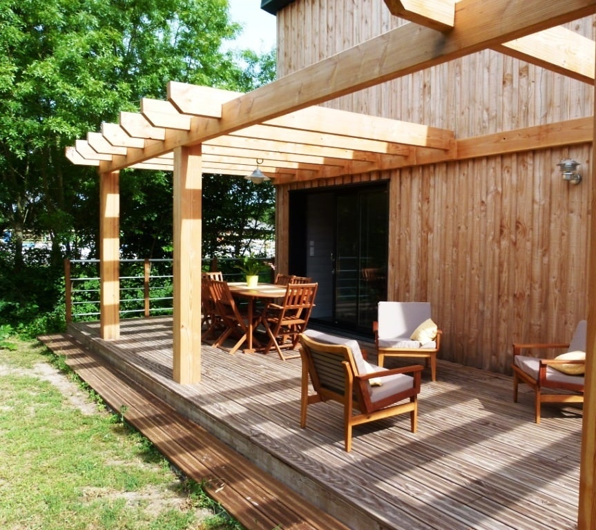 A wooden terrace showcases a spacious outdoor seating area with two armchairs and a round dining table surrounded by four chairs. Natural light filters through the wooden beams of the pergola, and the lush greenery in the background adds a serene touch.