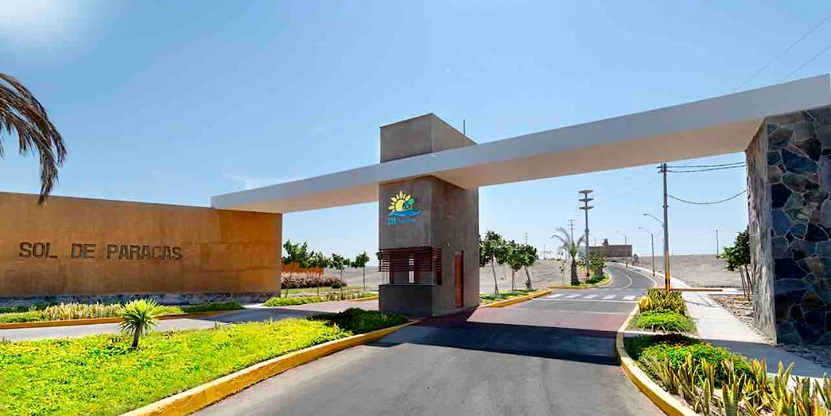 An entrance gate marked with 'Sol de Paracas' welcomes visitors. The bright blue sky shines overhead, while landscaped gardens line the pathway leading in. A paved road extends beyond the gate, flanked by low greenery and telephone poles in the distance.