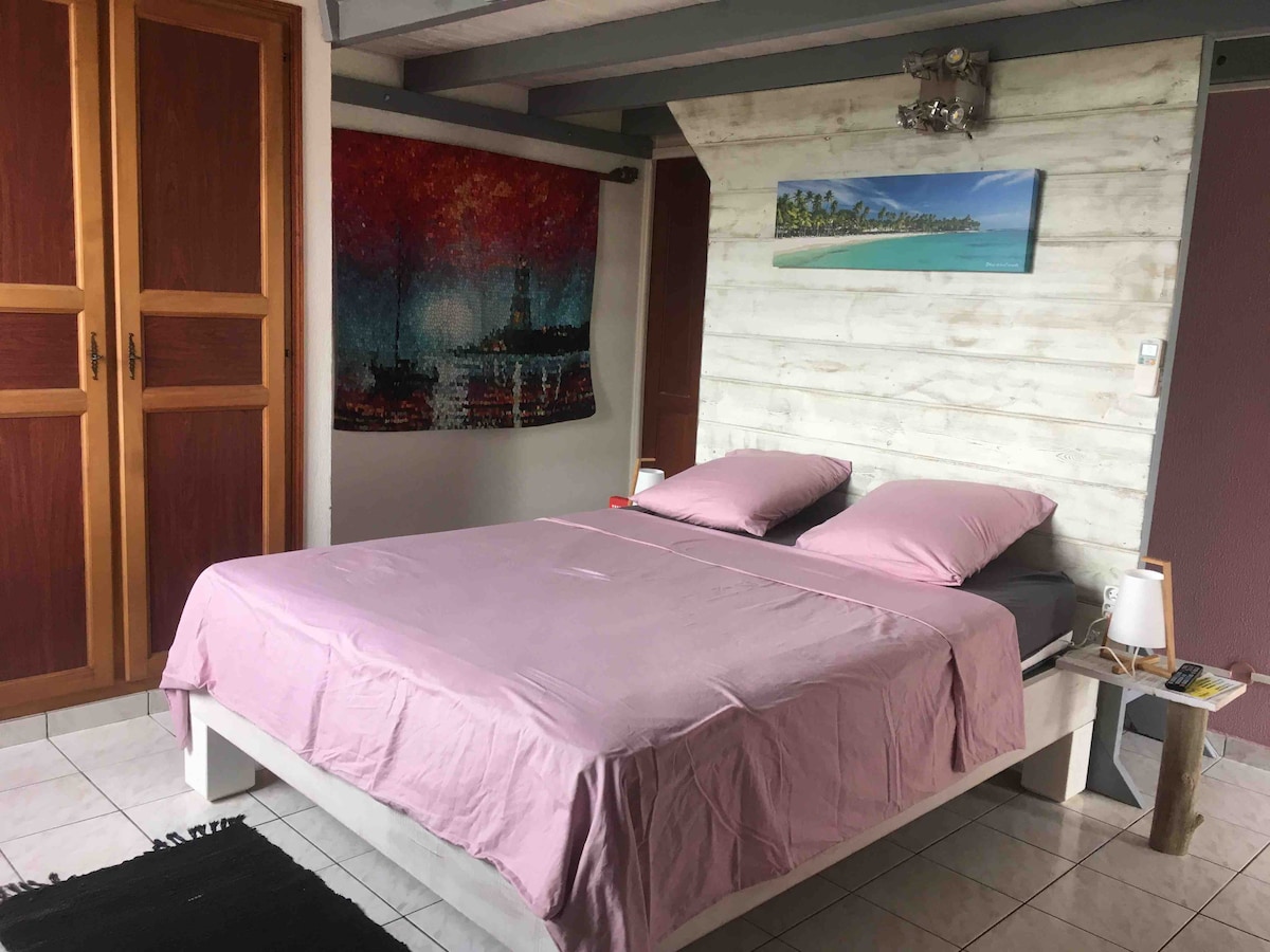 A spacious sleeping area features a large bed with light pink linens. The room is complemented by a colorful wall hanging and framed beach artwork above the bed. A compact bedside table and lamp add functionality, while the tiled floor provides a neutral base.