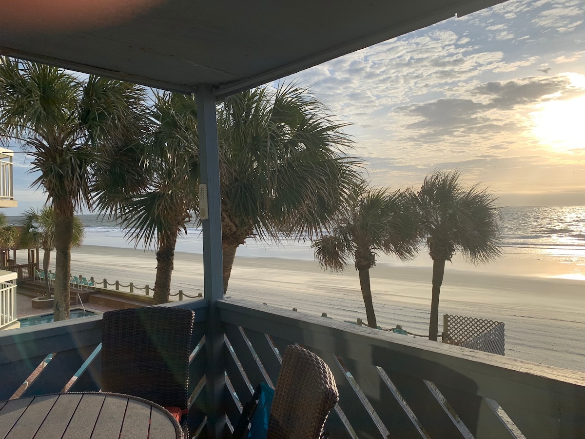 A cozy balcony setting overlooks a sandy beach framed by tall palm trees. The sun sets on the horizon, casting a warm glow over the water. A round table with wicker chairs is visible, enhancing the outdoor experience.